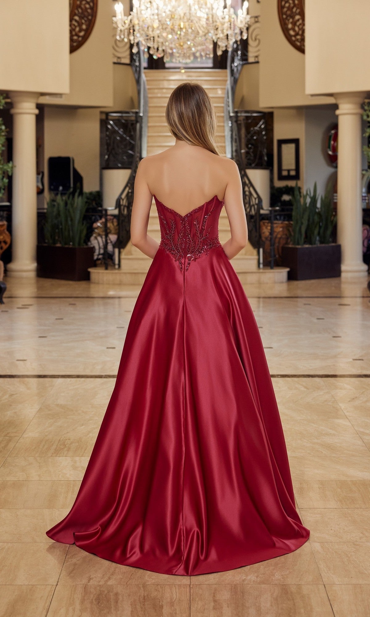 A woman wearing the Nox Anabel Y1798 long prom dress, featuring a strapless satin design and embellished corset bodice, stands with her back to the camera in an elegant foyer with marble floors and a chandelier.